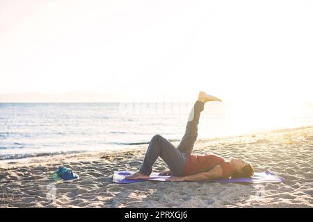 Femme âgée à la plage, sur un tapis de yoga, en train de faire de l'exercice. Lever les jambes. Banque D'Images