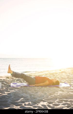 Femme âgée à la plage, sur un tapis de yoga, en train de faire de l'exercice. Lever deux fois la jambe. Banque D'Images