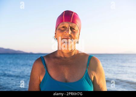 Femme âgée à la plage, debout au bord de la mer. Porter un maillot de bain bleu et un bonnet de bain rose, prêt à nager Banque D'Images