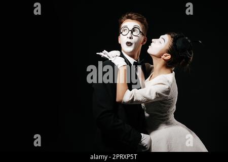 Portrait of couple mime sur fond noir. femme en robe blanche kissing man in tuxedo et verres Banque D'Images