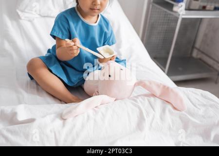 vue rognée de fille dans la robe d'hôpital nourrissant le lapin jouet avec le yogourt sur le lit dans la clinique, image de stock Banque D'Images