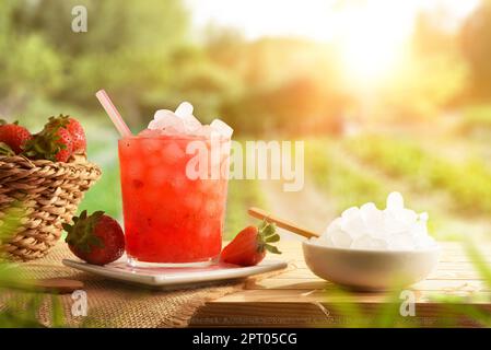Verre avec boisson naturelle de fraise avec beaucoup de glace et panier avec fruits sur une table en bois dans un verger de fraise dans la nature. Vue avant. Horizonta Banque D'Images