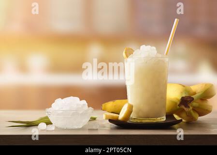 Deux verres avec boisson glacée à la banane sur une table en bois pleine de glace pilée et de fruits en tranches autour et fenêtre ensoleillée en arrière-plan. Vue avant. Horizon Banque D'Images
