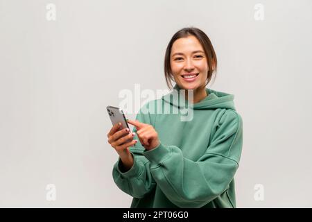 Femme de brunette asiatique satisfaite tenant un smartphone entre les mains, discutant avec des amis ou faisant des commandes de nourriture, services en ligne. Studio d'intérieur isolé sur fond blanc Banque D'Images