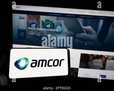 Personne tenant un téléphone portable avec le logo de la société d'emballage Amcor plc sur l'écran en face de la page Web d'affaires. Mise au point sur l'affichage du téléphone. Banque D'Images