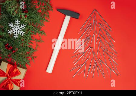 Clous longs et marteau, disposés en forme d'arbre de Noël avec branches d'épinette et boîte cadeau sur fond rouge.Pose à plat Banque D'Images