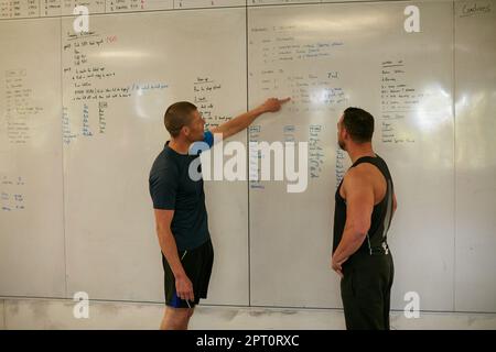 Visée IM pour le point numéro 1. deux jeunes hommes regardant un tableau blanc en se tenant dans la salle de sport Banque D'Images