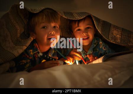 Maman dit que la lumière garde les mauvaises choses loin. Portrait de deux frères allongé sous leur couverture colorante en images avec une lampe de poche Banque D'Images