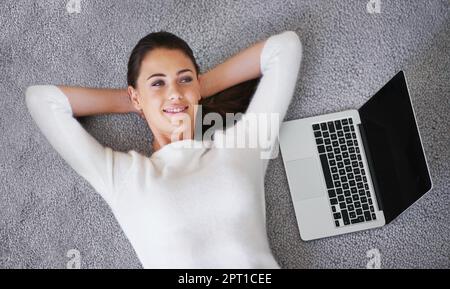 Si facile il fonctionne presque lui-même. Une jeune femme couché sur un tapis à côté d'un ordinateur portable Banque D'Images