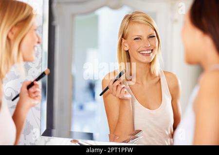 Sa peau est radieusement belle. une jeune femme appliquant le maquillage devant un miroir comme son ami se tient près Banque D'Images