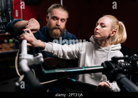 Professionnel barbu de vélo réparateur ayant une conversation avec la cliente blonde, parlant du problème de vélo détecté pendant le diagnostic Banque D'Images