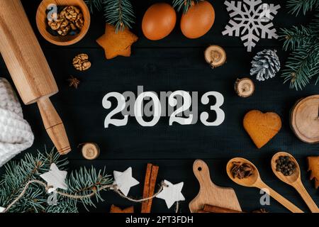 Les numéros 2023 en bois blanc se trouvent sur une table en bois noir avec des branches d'arbre de Noël, des accessoires de cuisson et des ingrédients. Joyeux Noël et bon N Banque D'Images