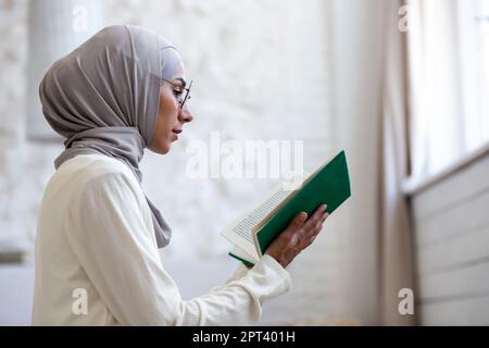 Gros plan. Une jeune femme dans un hijab et des lunettes, tenant un livre vert Saint dans ses mains, a plié ses mains et prie ensemble. Banque D'Images