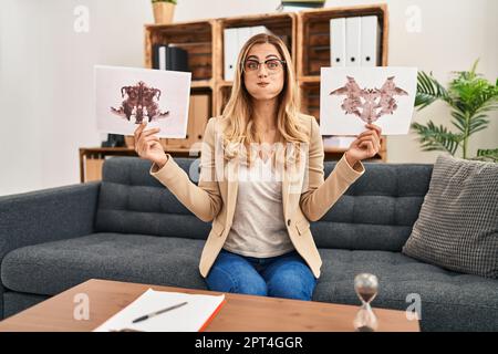 Jeune femme blonde thérapeute tenant rorschach test puffing joues avec visage drôle. bouche gonflée avec de l'air, attrapant l'air. Banque D'Images