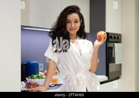 Bonne jeune femme brune attrayante femme au foyer tenant la pomme dans les mains souriant tout en regardant l'appareil photo debout dans la cuisine. Jolie femme de race blanche Banque D'Images