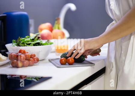 Gros plan des mains de femmes préparant le petit déjeuner dans la cuisine. Femme en train de trancher des légumes frais concombres et tomates sur planche à découper dans l'appartement Banque D'Images