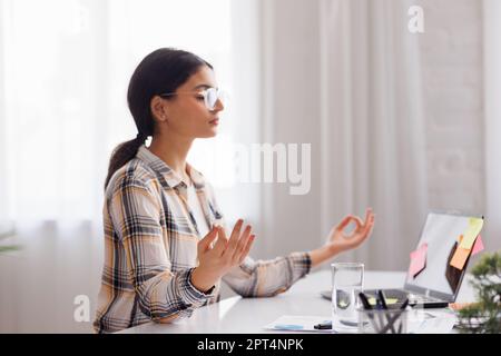 Gérer le stress sur le lieu de travail. Une étudiante hindoue médite et se détend pendant une journée bien remplie en étant assise sur son lieu de travail dans un bureau à l'intérieur Banque D'Images