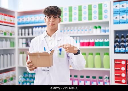 Adolescent hispanique travaillant à pharmacie drugstore tenant boîte avec des pilules sourire heureux pointant avec la main et le doigt Banque D'Images