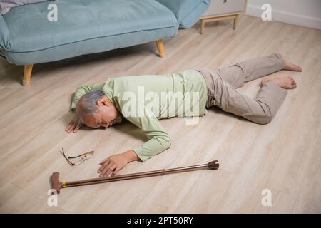 Homme asiatique âgé tombant couché sur le sol à la maison seul avec un bâton de marche en bois dans le salon, homme âgé maux de tête après la chute, soins de santé Banque D'Images