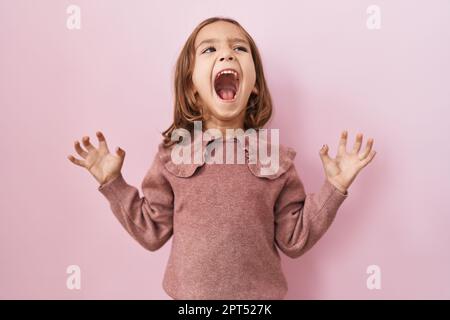 Petite fille hispanique debout sur fond rose fou et fou criant et hurlant avec l'expression agressive et les bras levés. concept de frustration. Banque D'Images