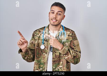 Jeune médecin hispanique portant un uniforme militaire de camouflage souriant et regardant la caméra pointant avec deux mains et des doigts sur le côté. Banque D'Images