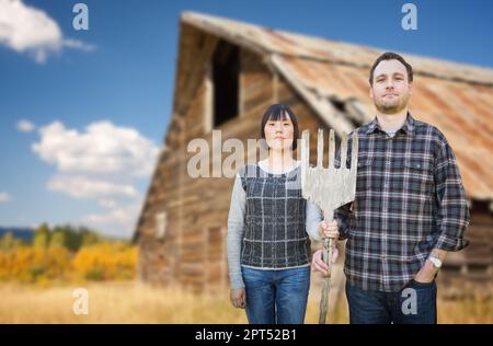 Jeune couple biracial chinois et caucasien tenant une fourche en bois en face de la Grange rustique dans le pays. Banque D'Images