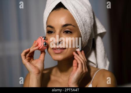 Gros plan de la jeune femme attirante caucasienne avec une serviette sur la tête en utilisant un appareil de virage facial à micro-courant à la maison. Appareil de beauté. Belle femme heureuse Banque D'Images