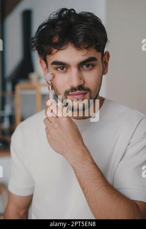 Portrait d'un homme utilisant le rouleau de jade massant son visage avec pour une peau plus tonifiée et brillante. Beau jeune homme pratiquant dans l'auto-soin anti-âge Banque D'Images