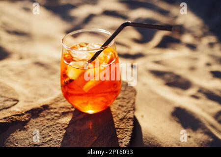 Tranche d'orange et apéritif spritz cocktail sur une pierre sur fond d'une plage de sable. Verre à l'eau minérale pétillante aux agrumes et à la paille. Banque D'Images