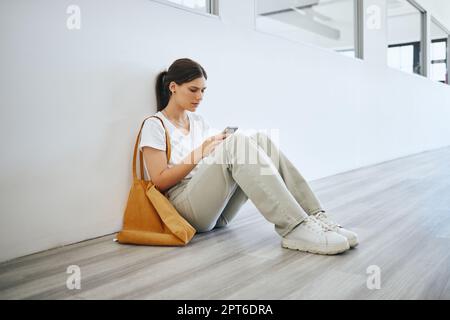 Triste femme universitaire, étudiante seule sur le sol dans le couloir après la classe ou la conférence et la navigation sur les médias sociaux sur smartphone. Dépression universitaire, s Banque D'Images