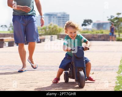 HES né pour être un garçon de biker. Un jeune garçon heureux de conduire est moto jouet dehors tandis que son père court après lui de derrière Banque D'Images