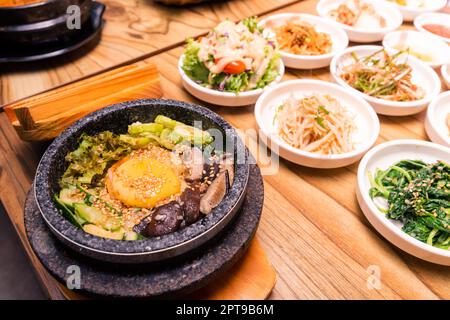 Bibimbap dans un bol en pierre chauffée noir, plat traditionnel coréen - bibimbap mélange de riz avec des légumes comprend le boeuf et l'œuf frit et les cornichons mis sur le tabl Banque D'Images