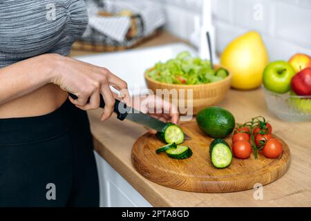 Concept de saine alimentation. Gros plan de la jeune femme afro-américaine dans un vêtement de sport, debout à la maison dans la cuisine en coupant le concombre tout en préparant de la salade fraîche utile, souriant heureux Banque D'Images