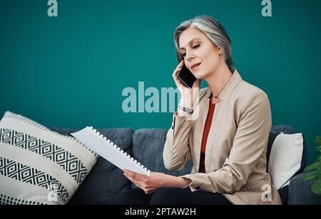 Je passe en revue les documents au fur et à mesure que nous parlons. une femme d'affaires attirante et mûre assise seule et utilisant son téléphone portable tout en lisant la paperasse dans son ho Banque D'Images