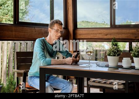 Un homme souriant d'âge moyen boit du vin assis sur la véranda de sa maison de campagne et utilisant un smartphone Banque D'Images