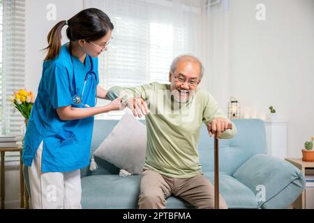 Une jeune femme aide à soutenir les patients orthopédiques pour se lever avec la canne à pied à la maison, une infirmière de soins aide à soutenir l'homme handicapé senior pour se lever avec Banque D'Images