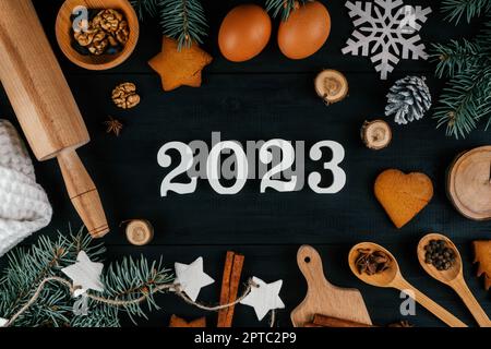 Les numéros 2023 en bois blanc se trouvent sur une table en bois noir avec des branches d'arbre de Noël, des accessoires de cuisson et des ingrédients. Joyeux Noël et bon N Banque D'Images