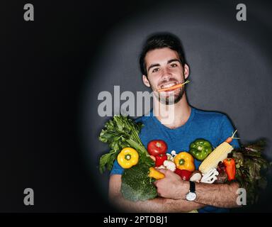 Sain à l'extérieur commence à l'intérieur. Portrait en studio d'un jeune homme portant une quantité de légumes sains sur un fond sombre Banque D'Images