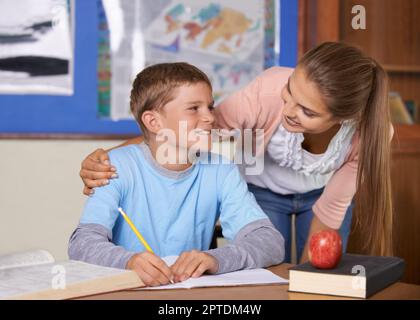 Je prends du temps pour tous mes étudiants. Un jeune enseignant de soutien se penchait sur son élève pendant qu'il écrit dans un livre Banque D'Images