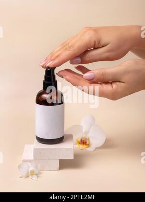 Une femme pompe à la main du liquide sur une main courte d'une bouteille cosmétique près des fleurs d'orchidées sur beige gros plan, maquette d'étiquette. Kit de produits de beauté pour le soin de la peau Banque D'Images