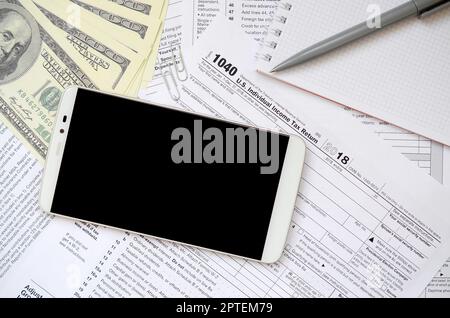 Composition d'articles se trouvant sur le formulaire fiscal 1040. Dollar bills, stylo, smartphone, trombone et le bloc-notes. Nettoyer l'écran noir pour smartphone text placeme Banque D'Images