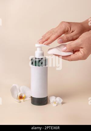 Une femme pompe à la main du liquide sur un tampon démaquillant d'une bouteille cosmétique près des fleurs d'orchidées sur beige gros plan, maquette d'étiquette. Produit de beauté pour les soins de la peau Banque D'Images