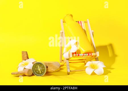 Chaise-terrasse miniature avec bouteille de crème solaire, boussole, avion en bois et fleurs de plumeria sur fond jaune Banque D'Images