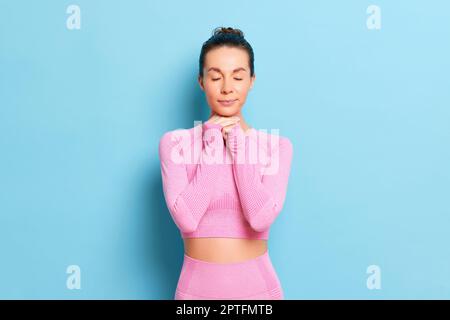 Portrait d'une jeune femme heureuse aux yeux fermés, elle porte un costume de yoga rose tenant les deux mains sous son menton, posant sur fond bleu studio, cop Banque D'Images