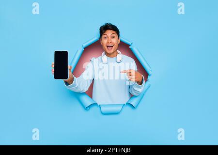 Jeune homme souriant modèle en chemise bleu clair debout dans le trou au centre de fond bleu, homme avec le téléphone dans sa main pointe son doigt vers t Banque D'Images