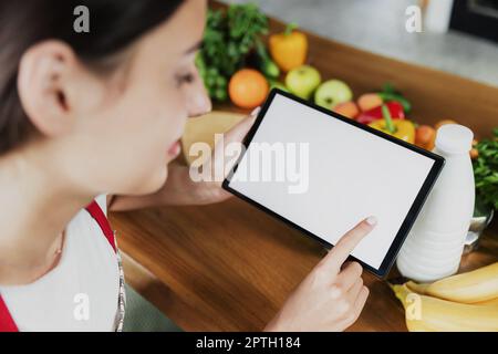 Jeune femme utilisant un smartphone, fille mains tenant et cliquant sur la tablette avec vide écran blanc maquette. Plats frais sur la table de la cuisine. Cuisson Banque D'Images