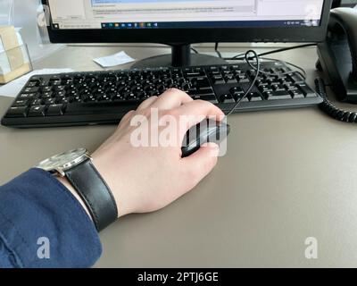La main d'un homme avec une chemise et une horloge tient une souris d'ordinateur sur la table de bureau avec un ordinateur avec un clavier. Travail d'entreprise. Banque D'Images