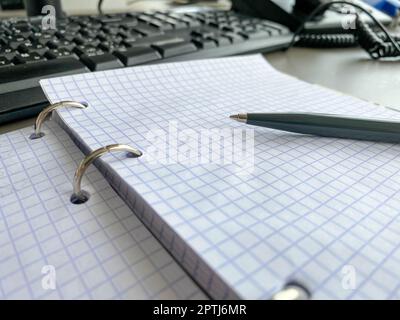 Un stylo à écrire repose sur un bloc-notes avec des feuilles de papier carrées sur un bureau de travail avec de la papeterie dans un bureau d'affaires. Banque D'Images