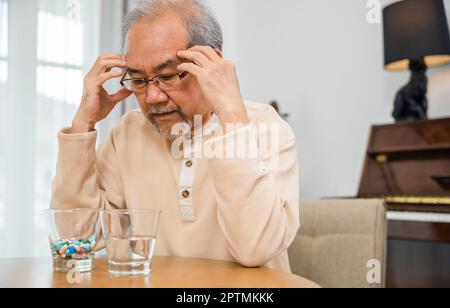 Asiatique senior vieux homme gris cheveux triste assis dans la chaise main tenir la tête avec de nombreux médicaments et pilules pour prendre, homme âgé malade ont mal de tête regardant Banque D'Images