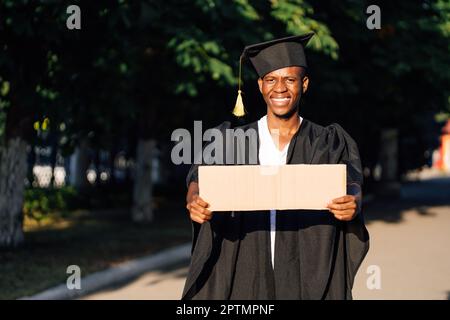 Portrait d'un homme noir heureux debout avec une affiche en carton vide dans la rue à la recherche d'un travail, espace libre de copie. Étudiant diplômé d'une université ou d'un collège Banque D'Images